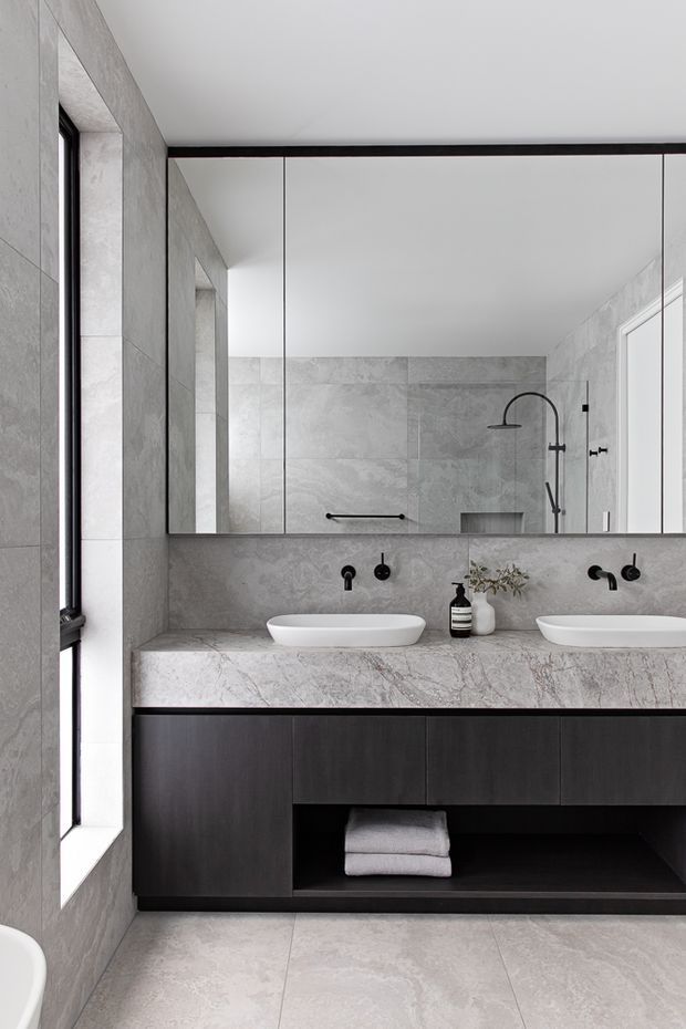Contemporary double-vanity bathroom featuring large-format grey stone-look wall and floor tiles, a pale veined stone benchtop, frameless mirrored cabinetry, matte black wall-mounted tapware, and dark timber or black vanity joinery. A walk-in shower is reflected in the mirror, and a freestanding bath is partially visible at the lower left.