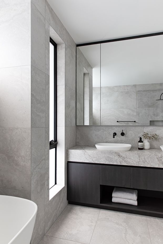 A contemporary bathroom with large-format grey stone-look wall and floor tiles, a freestanding white bathtub, frameless mirrored cabinetry, a white vessel basin, dark timber vanity cabinetry, and black fixtures. A narrow black-framed window provides natural light.