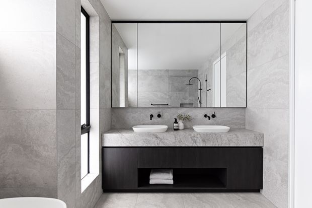 Contemporary bathroom featuring a double vanity with white vessel basins, a dark timber-toned floating cabinet, pale grey stone-look wall and floor tiles, matte black tapware, and a large mirrored cabinet above the vanity. A shower area is visible in the reflection.