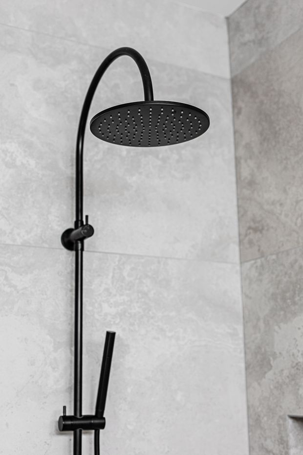 Close-up of a matte black rainfall shower system with curved riser, round overhead showerhead, handheld shower wand, and controls mounted against large light grey stone-look wall tiles in a bathroom.