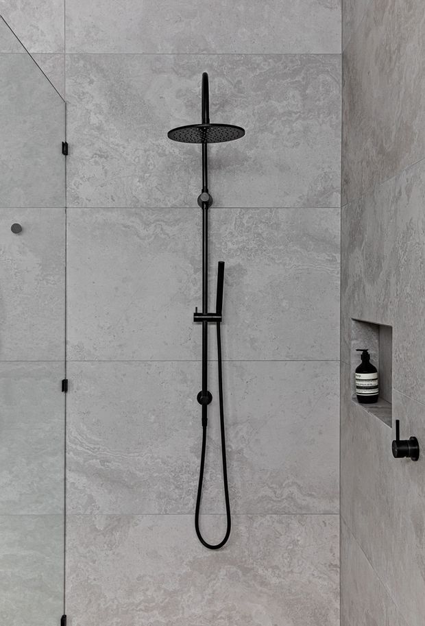 A minimalist walk-in shower clad in large-format pale grey stone-look tiles. Matte black fixtures include a round overhead rain shower, handheld shower on a vertical rail, hose, controls, and a glass shower door. A recessed wall niche holds a dark soap dispenser, creating a clean, monochromatic bathroom interior.