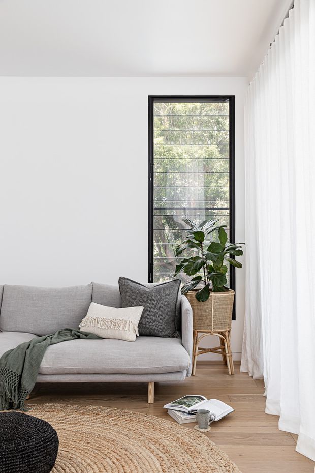 Bright, calm living room with a light grey sofa, textured cushions, a woven throw, a large round natural-fibre rug, and a potted plant beside a tall black-framed louvered window. White walls and sheer floor-to-ceiling curtains create an airy interior, with light timber flooring and a small stack of books and cup on the floor.