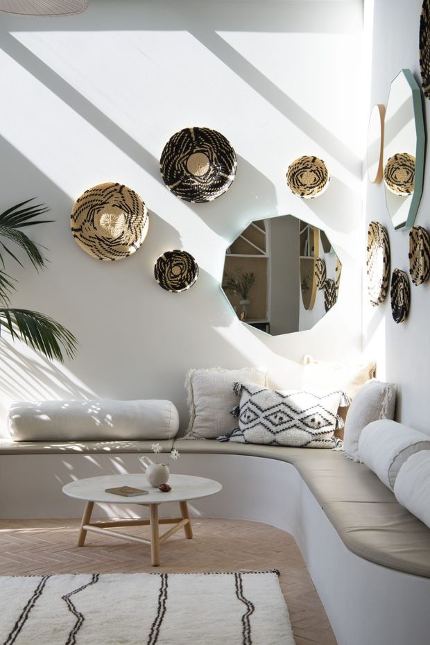 Bright, sunlit living room with a built-in curved white sectional sofa, white walls, woven black-and-natural wall baskets, an organic-shaped mirror, a small round timber coffee table, and a neutral patterned rug. Strong diagonal sunlight creates a warm, airy atmosphere, with palm foliage visible at the edge of the room.