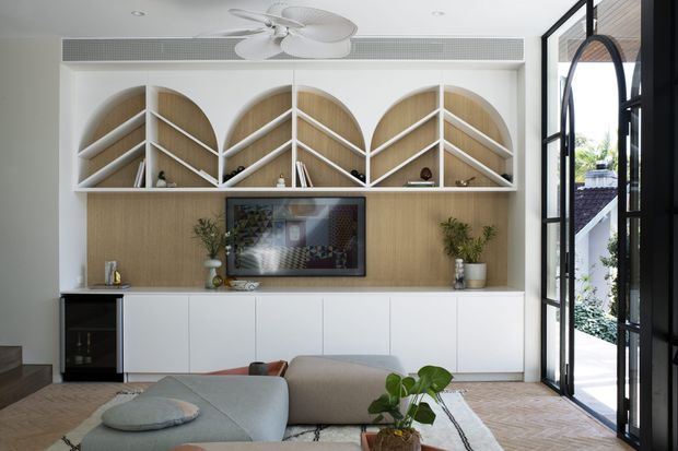 Bright contemporary living room with a white built-in media cabinet and open display shelving featuring arched, timber-lined niches. A television, indoor plants, soft seating, patterned rug, sculptural ceiling fan, and full-height black-framed glazed doors create a refined, airy interior.