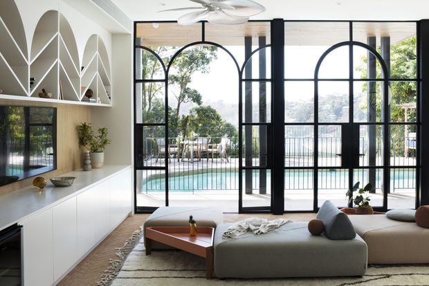Bright contemporary living room with a large built-in media wall and sculptural arched shelving, a low modular sectional sofa, and extensive black-framed arched glazing opening to a pool terrace. The room has light timber flooring, pale neutral joinery, and a ceiling fan, with leafy views and a hillside cityscape beyond.