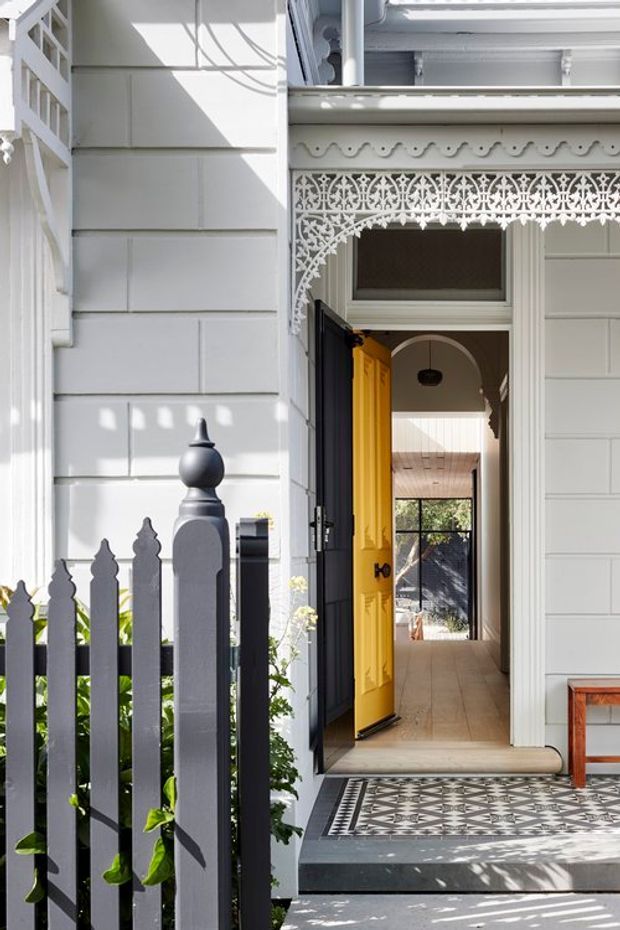 A bright exterior view of a heritage-style residential entryway with white weatherboard cladding, ornate lacework and moulded trim, a decorative picket fence, and a vivid yellow front door opened into a tiled hallway. Patterned encaustic-style tiles form the front porch, while a timber-lined interior and garden planting are visible beyond the entry.