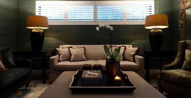 A compact, richly layered living room with dark textured wall panelling, a wide horizontal bank of white plantation shutters, and a symmetrical seating arrangement. A neutral upholstered sofa sits between two black table lamps on dark side tables, facing a large upholstered ottoman and black tray coffee table. A dark lounge chair and patterned armchair frame the composition, while an orchid and candle add restrained decorative detail. The room has a moody, intimate, refined atmosphere with strong symmetry and warm lamp light.