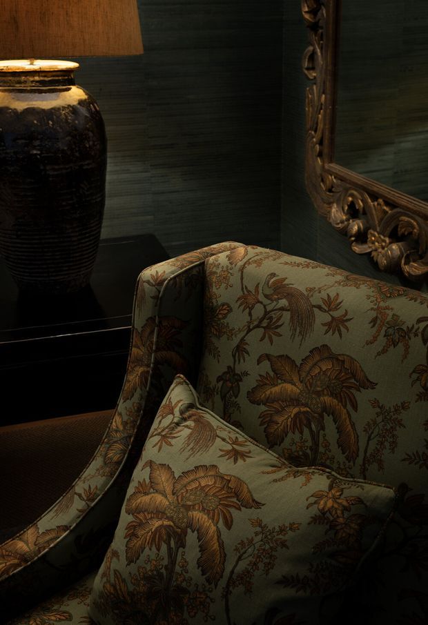 Close-up, moody interior vignette featuring a patterned upholstered armchair in muted sage green with botanical motifs, beside a glossy dark ceramic table lamp and a dark console. An ornate carved gilt mirror and dark textured wall finish create a richly layered, traditional atmosphere.