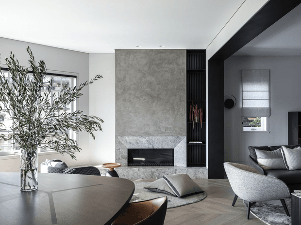 Refined contemporary open-plan living and dining room with a large round dining table in the foreground, sculptural chairs, a grey sectional sofa, and a central textured stone fireplace. Dark built-in shelving and a broad black structural opening define the transition to the adjoining sitting area. Neutral finishes, herringbone timber flooring, soft furnishings, and abundant natural light create a calm, sophisticated interior.