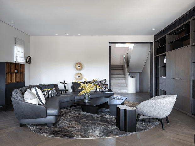 A refined contemporary living room with a large charcoal sectional sofa, sculptural lounge chair, black coffee table, patterned area rug, and dark built-in shelving and cabinetry. A staircase is visible through a wide opening at the rear, while neutral walls, recessed ceiling lights, and warm timber flooring create a sophisticated, monochromatic interior.