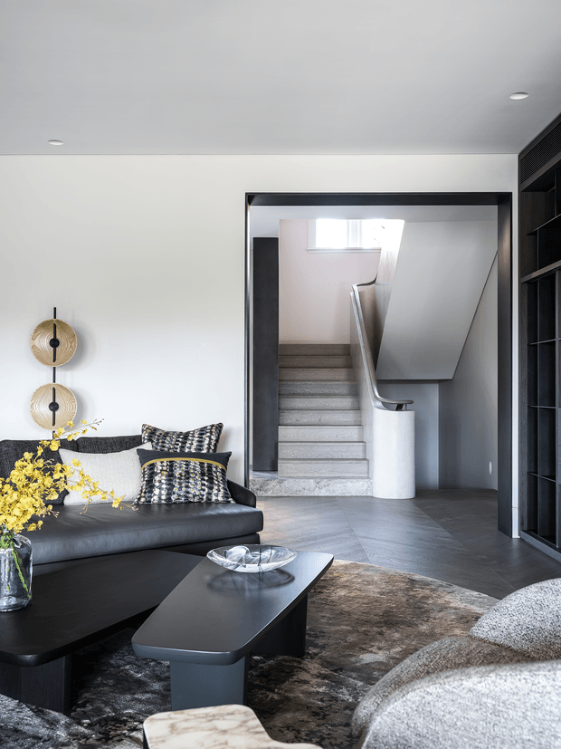 Refined contemporary living room with a black leather sofa, sculptural dark coffee tables, textured cushions, a large patterned rug, and a bright floral arrangement. A wide opening reveals a modern staircase with pale stone or concrete treads, a curved white balustrade, and dark framing. Built-in dark shelving and ventilation details line the right side, while minimalist wall-mounted decorative discs add visual interest.