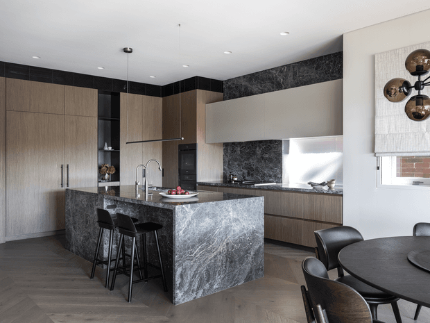 A sophisticated contemporary kitchen and dining area featuring full-height light timber cabinetry, a large dark veined stone island with waterfall ends, matching stone splashback accents, matte black appliances and tapware, and dark herringbone timber flooring. Black bar stools sit at the island, while a round dining table and chairs occupy the foreground. A sculptural dark wall light and textured roller blind add decorative detail.