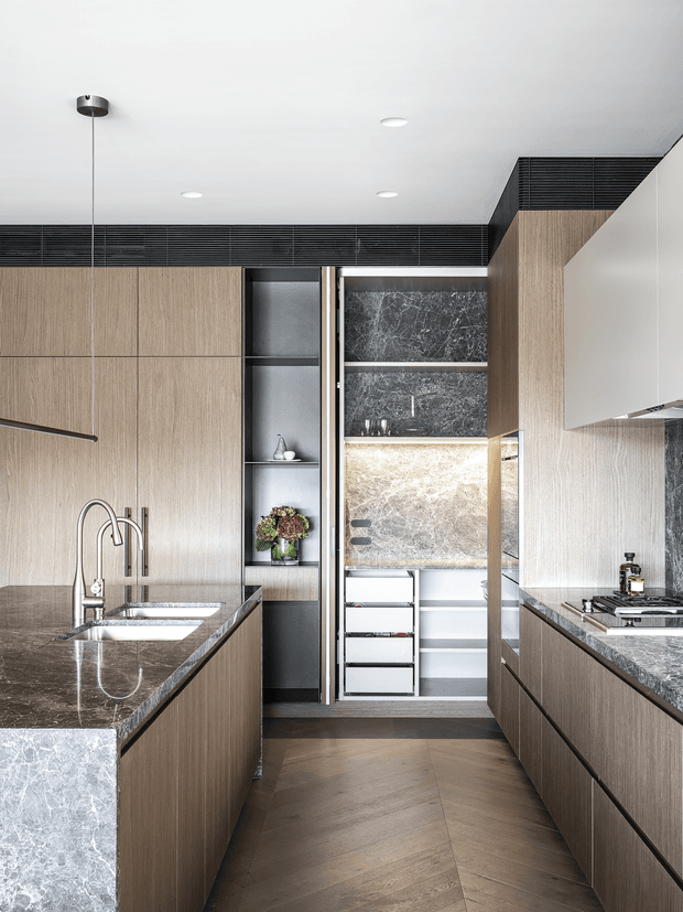 A refined contemporary kitchen with full-height light timber cabinetry, dark veined stone benchtops, an island with integrated double sink and sculptural tapware, concealed appliances, open display shelving, and a compact walk-in pantry or secondary preparation area at the rear. The space has recessed ceiling lights, dark linear ceiling detailing, and warm herringbone timber flooring.