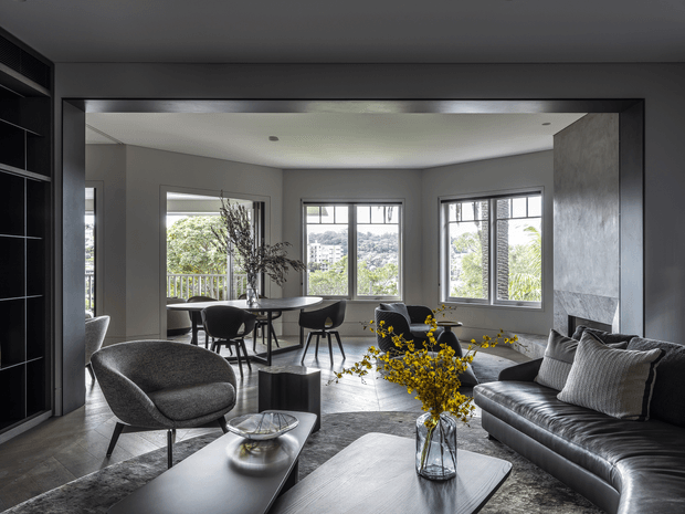 A refined contemporary living and dining area viewed from the living room. The space features a dark leather sofa, sculptural lounge chair, coffee tables, a round dining table with chairs, large black-framed windows, grey walls, dark built-in joinery, and a textured stone-like feature wall with a broad horizontal fireplace or wall opening. Yellow flowering branches provide a bright accent against the muted interior.