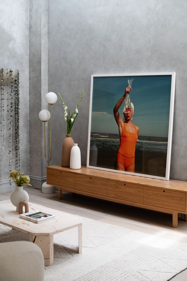 A refined living room with a warm timber low-profile media console, large framed artwork, sculptural floor lamp, ceramic vases, a pale textured rug, and a light stone coffee table. The room features cool grey textured walls balanced by warm wood and soft neutral furnishings.