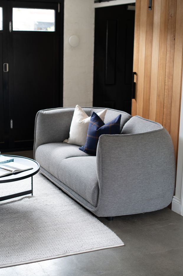 A compact, refined living room featuring a rounded light-grey upholstered sofa with navy and cream cushions, a black-framed circular coffee table, a textured pale rug, dark painted doors, and warm vertical timber wall panelling. The space has a contemporary, balanced palette with strong black, grey, white, and natural wood contrasts.