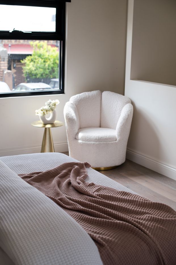 A softly lit bedroom corner with a neatly made bed in the foreground, a plush cream boucle armchair, and a small round gold side table holding a vase of white flowers. The room has warm off-white walls with simple trim, pale timber flooring, and a black-framed window overlooking an urban street. A neutral taupe knitted throw adds texture across the bed.