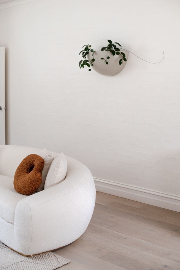 A calm, minimalist interior corner featuring a sculptural white boucle armchair with a rust-coloured cushion, pale timber flooring, and a white-painted brick wall. A wall-mounted rounded planter with trailing greenery adds a natural accent, while simple white trim and a partially visible doorway frame the space.