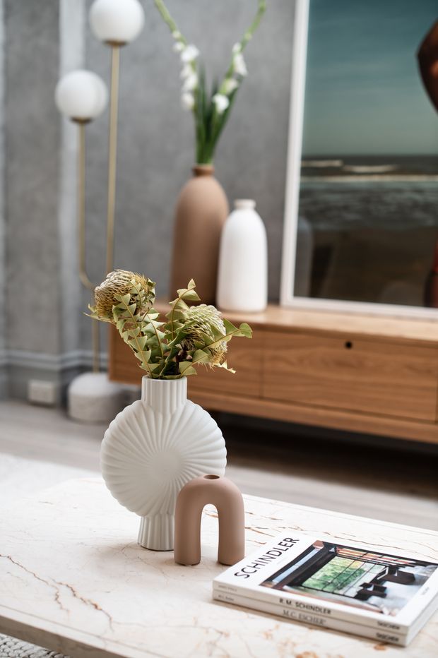 Styled living room vignette featuring a veined stone coffee table with sculptural ceramic vases and dried botanical arrangements in the foreground. A timber media console, large television, floor lamps, and a textured grey wall create a warm contemporary interior setting.