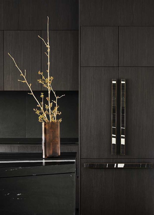 Close-up of a sophisticated dark-toned built-in kitchen or cabinetry wall. Vertical black-stained wood-grain panels form tall cupboard doors with long slim metallic handles, paired with glossy black stone or glass surfaces and a dark reflective splashback. A copper-toned vase with delicate branches provides a warm natural accent.