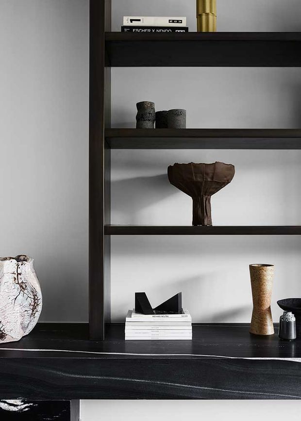 Close-up interior detail of a dark timber or dark-stained built-in shelving unit against a light wall, styled with ceramic vessels, sculptural pottery, books, and a brass-toned decorative object. The composition has a refined, moody, minimalist feel with strong horizontal shelving and contrasting dark surfaces.