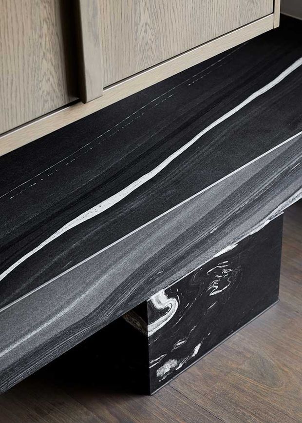 Close-up detail of a dark, heavily veined stone benchtop or counter with a contrasting white mineral vein, paired with a dark stone support and light wood-grain cabinetry. A timber-look floor is visible below.