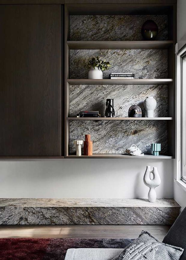 Close-up interior view of a refined built-in storage and display unit beside a window. Dark timber cabinetry frames open shelves lined with dramatic grey veined stone, matching the stone countertop and raised ledge below. Minimal decorative objects, a neutral wall, timber flooring, and the edge of a sofa create a sophisticated living-room or study setting.