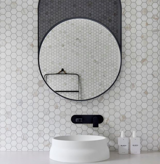 Close-up view of a contemporary bathroom vanity featuring a white vessel basin on a pale countertop, black wall-mounted tapware, and a large round black-framed mirror. The wall is covered in white and light grey hexagonal mosaic tiles, with matching dark hexagonal tiles visible inside the mirror reflection. Minimal white bathroom accessories sit beside the basin.