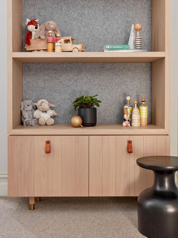 A styled built-in timber storage unit with two open display shelves backed in grey felt and lower closed cupboards with leather pull handles. The shelves hold toys, books, small decorative objects, and a potted plant, while a sculptural black stool sits in the foreground. The room has a warm, playful, carefully curated contemporary feel.