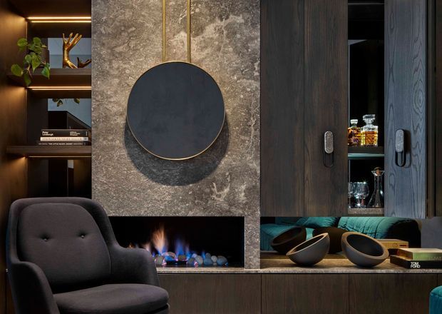 A moody, refined built-in living room or media joinery wall featuring dark timber cabinetry, open shelving with decorative objects and books, a stone-clad fireplace surround, a round dark feature panel, and a linear gas fireplace. A sculptural dark armchair and decorative bowls sit in the foreground, with concealed warm LED lighting and a small drinks cabinet integrated into the joinery.