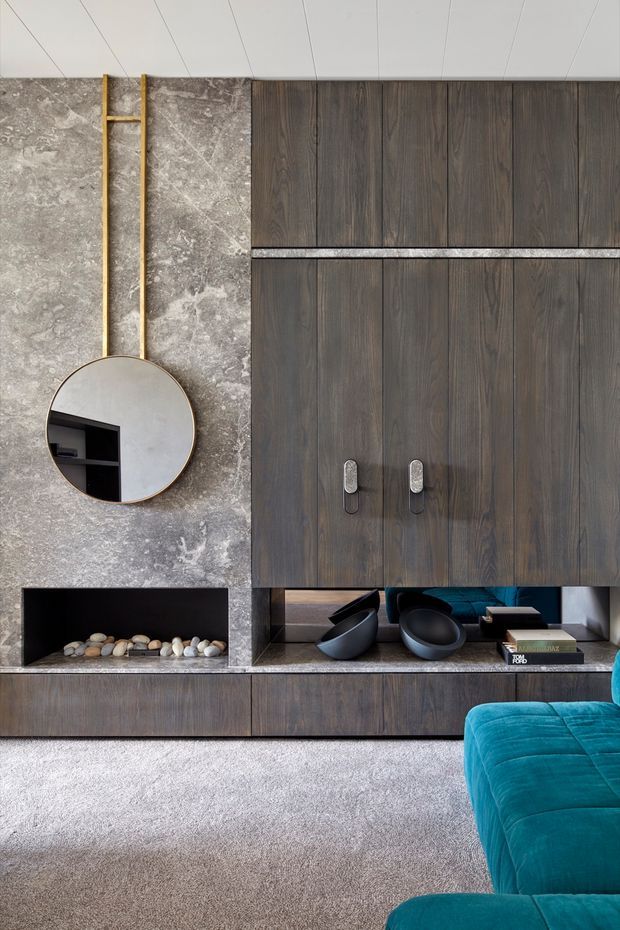 A refined contemporary living room media wall featuring dark timber cabinetry with integrated shelving, a grey stone-look feature wall, a slim linear fireplace filled with decorative stones, and a round wall mirror with a brass-toned frame. A vivid teal upholstered sofa occupies the foreground, while sculptural black bowls and books style the open shelving. The space has a restrained, luxurious palette with strong material contrast.