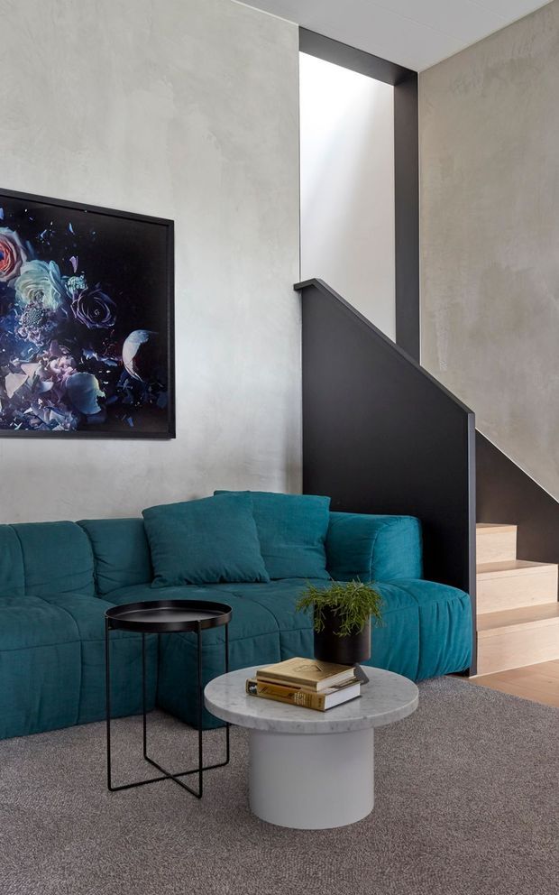 A contemporary living room with a deep teal sectional sofa, black metal side table, round grey stone coffee table, and framed artwork. The room features textured light grey walls, grey carpet, a dark sculptural stair balustrade, and pale timber stair treads leading upward.