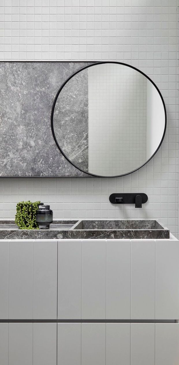 A close-up view of a contemporary bathroom vanity featuring a large round black-framed mirror, white small-format tiled walls, a grey veined stone backsplash and integrated double basin countertop, black wall-mounted tapware, and white vertically panelled vanity cabinetry. A small plant and dark bathroom accessories sit on the counter.