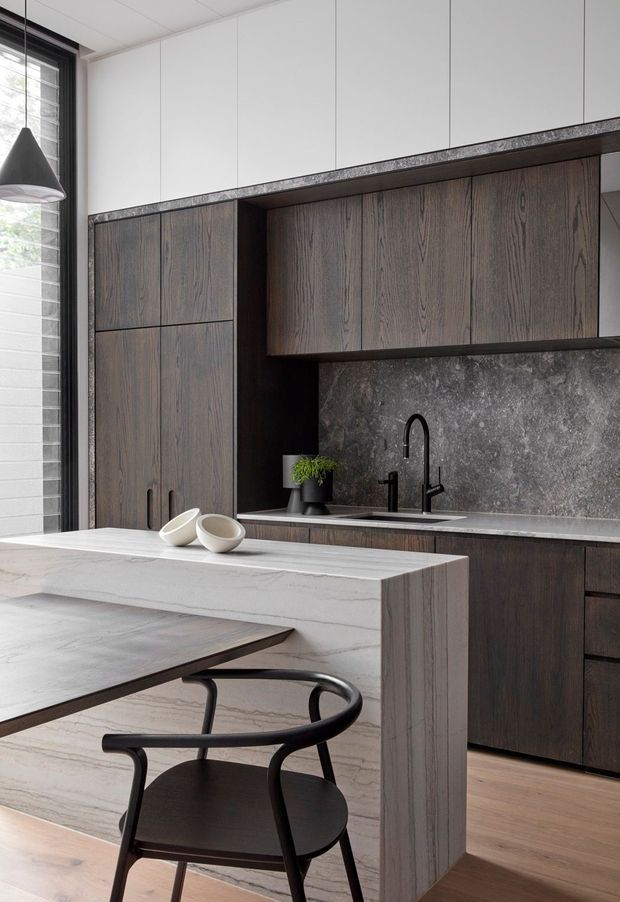 A refined contemporary kitchen featuring dark timber-grain cabinetry, a large pale stone island with waterfall edge, dark stone splashback, black tapware and a black sculptural dining chair. A tall window and pendant light provide natural light, while timber flooring adds warmth.