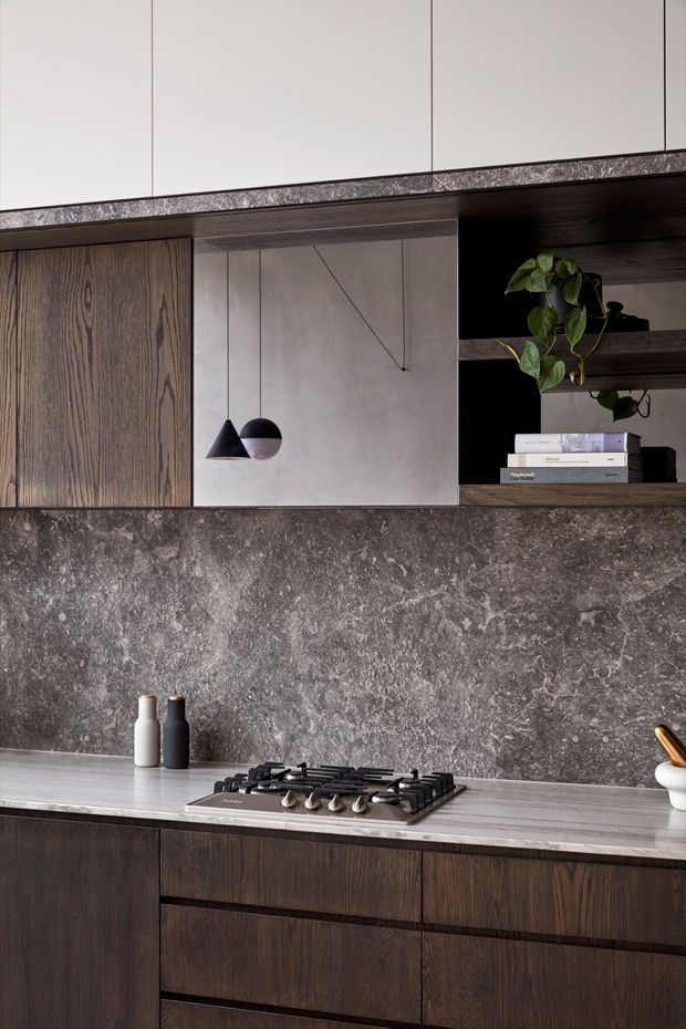 A refined contemporary kitchen featuring dark timber-grain cabinetry, a dramatic grey stone splashback and benchtop, integrated open shelving, and a central gas cooktop. Minimal pendant lights, neutral overhead cabinetry, and a trailing plant create a warm, sophisticated material palette.