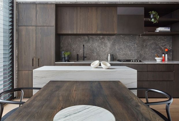 Contemporary kitchen and dining area with dark timber cabinetry, a grey stone backsplash and benchtops, a white stone island, and a dark timber dining table with curved black chairs. Vertical window blinds and open shelving add architectural detail, while restrained styling creates a refined, moody atmosphere.