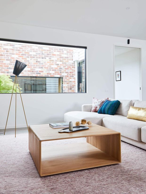 Bright contemporary living room with a large cream sectional sofa, pale timber open-base coffee table, soft textured rug, tripod floor lamp, and colourful cushions. A large internal window provides a view toward exposed brick and glazed architectural elements, while a white doorway and walls keep the room light and minimal.