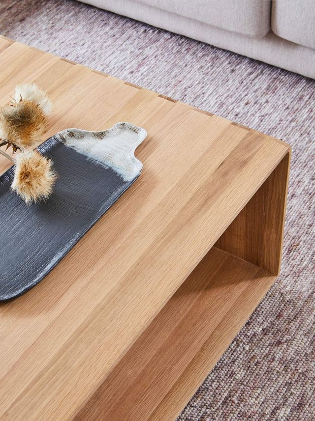 Close-up interior detail of a light oak timber coffee table with a layered open-shelf design, positioned on a textured neutral rug beside a pale upholstered sofa. A dark decorative tray with dried botanical accents sits on the tabletop.