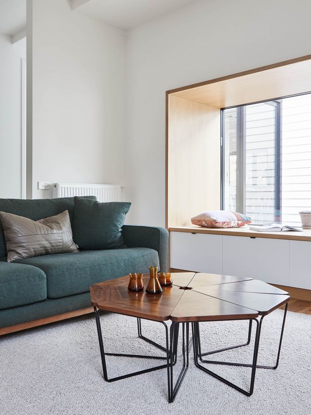Bright, contemporary living room with a teal upholstered sofa, patterned cushions, a geometric dark timber coffee table with black metal legs, and a light carpet. A built-in timber-lined window seat and white cabinetry sit beside large glazing, creating a clean, airy interior.