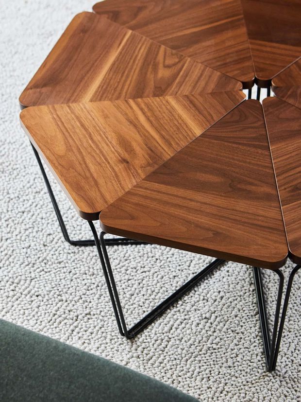 Close-up product photograph of a modular geometric coffee table made from multiple interlocking triangular and rounded-edge walnut-toned timber sections, supported by slim black metal hairpin-style legs. The table sits on a light textured carpet beside a dark green upholstered surface.