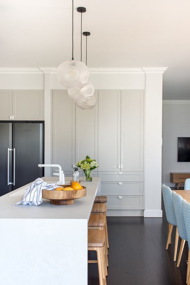 Bright, refined kitchen interior featuring a large pale island with integrated white sink and tap, warm timber stools, full-height panelled cabinetry in a soft grey-beige tone, dark flooring, and a black refrigerator. Sculptural white pendant lights hang above the island, while a dining area with blue upholstered chairs and a wall-mounted television is visible to the right.