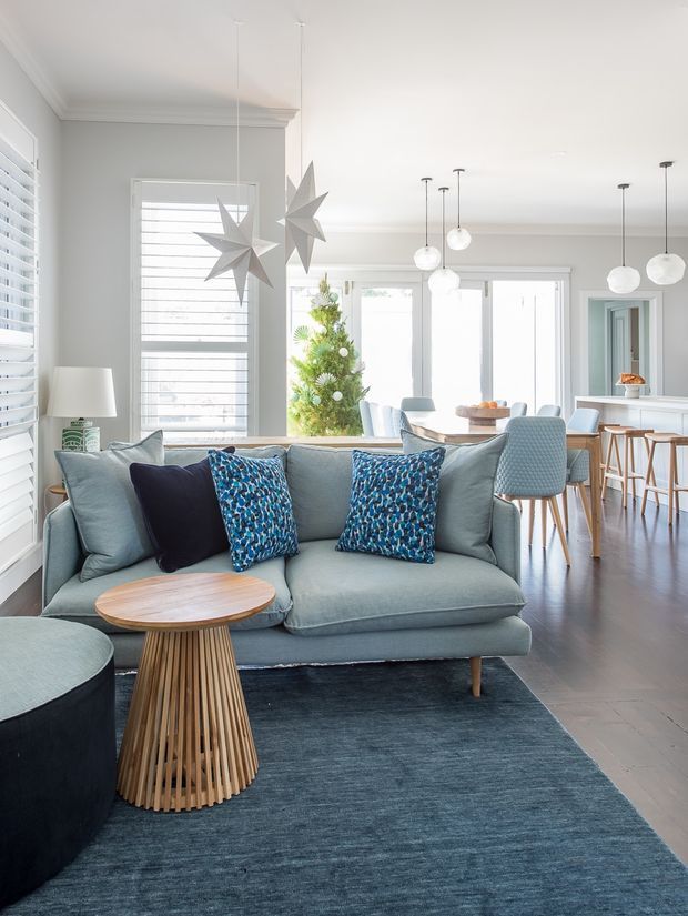 Bright open-plan living and dining area with a pale blue-grey sofa, patterned blue cushions, dark rug, and a sculptural timber coffee table. The room features white plantation shutters, light grey walls with decorative cornices, large glazed doors, multiple white pendant lights, a dining table, and a kitchen or bar area with stools in the background. A small decorated tree is visible near the glazing.