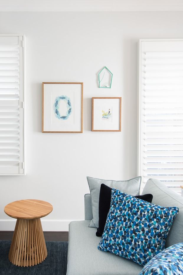 Bright, carefully styled living room corner with a pale upholstered sofa, layered blue patterned and dark cushions, a round slatted timber side table, framed artwork, a small decorative wall piece, white plantation shutters, and a dark rug. The room has a light, fresh palette with blue accents and natural timber detailing.