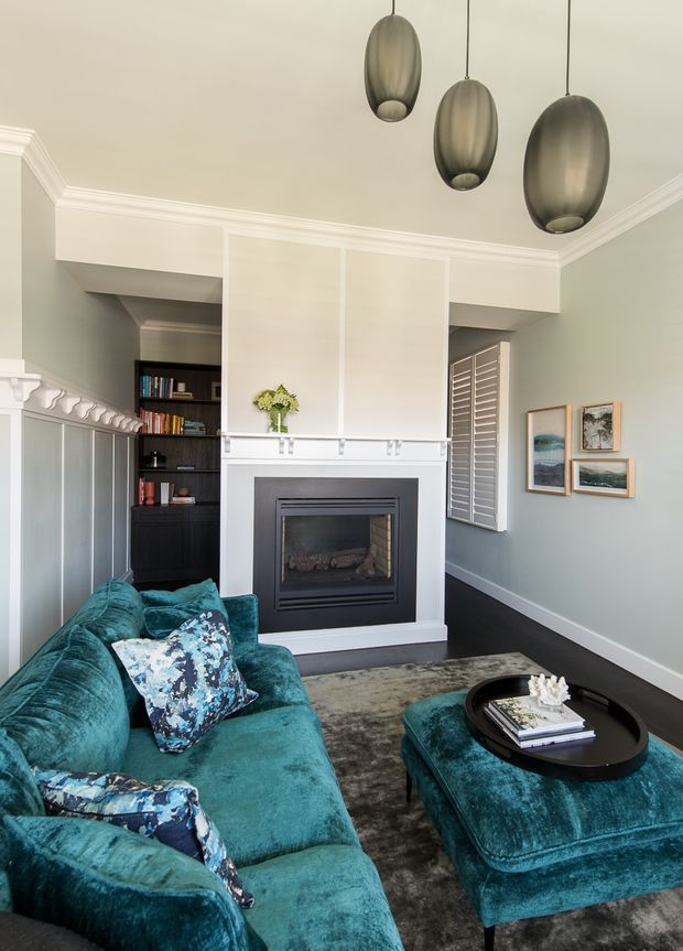 A refined living room with a black fireplace centred on a pale panelled chimney breast, dark built-in shelving, ornate white cornices and wall panelling, and plantation shutters. A deep teal velvet sofa and matching ottoman surround a black oval coffee table, with three sculptural pendant lights overhead and framed artwork on the muted green-grey walls.