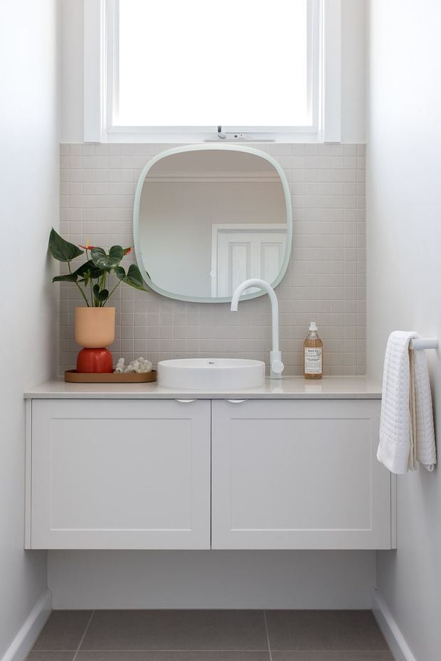 A bright, compact bathroom vanity with a floating white panelled cabinet, white vessel basin, sculptural white tapware, and a rounded rectangular mirror. The vanity is set against a pale grey tiled splashback beneath a large window, with a small plant, soap dispenser, and folded towels adding restrained decorative detail.