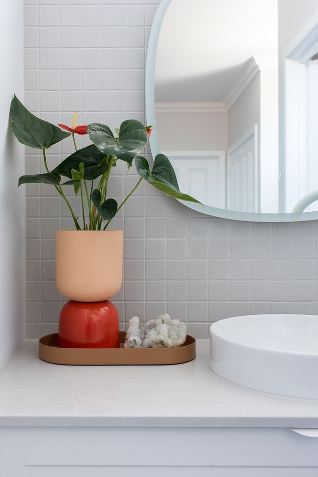 Close-up of a bright, neatly styled bathroom vanity. A white tiled wall with fine grout lines sits behind a large rounded-edge mirror, reflecting a white paneled interior door and ceiling trim. On the pale countertop is a warm terracotta-toned tray holding a peach ceramic planter with a leafy tropical plant, a glossy red rounded vessel, and a small pale mineral-like decorative object. The edge of a white basin is visible at right, with white painted vanity cabinetry below.