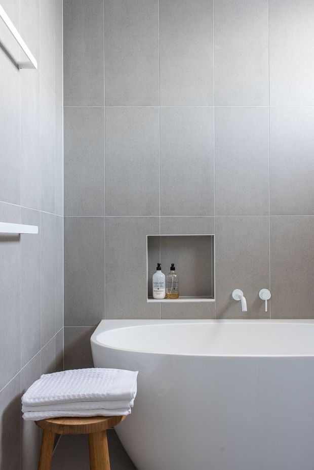 A minimalist bathroom with a freestanding white bathtub against large-format light grey tiled walls. A recessed wall niche holds bath products, with understated white wall-mounted tapware beside the tub. A small timber stool with folded white towels sits in the foreground, and a simple white shelf is visible on the left wall.