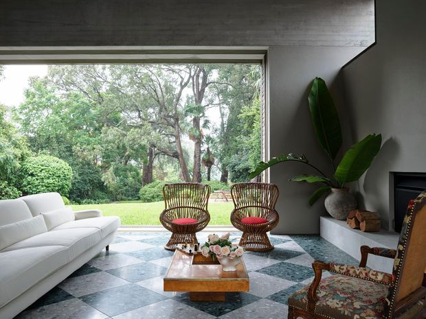 A refined living room with a large open picture window overlooking lush greenery. The room features a white sofa, two woven rattan lounge chairs with red cushions, an ornate upholstered armchair, a low wooden coffee table with flowers, a large potted plant, a dark fireplace surround, and a geometric checkerboard stone floor. The space combines indoor comfort with a strong connection to the garden.