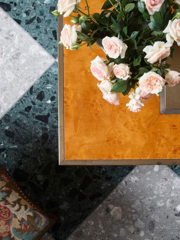 Top-down detail of a richly styled interior vignette featuring a polished orange-brown burl wood tabletop with a metal border, set over dark green and pale grey terrazzo flooring. A bouquet of pale pink and cream roses sits on the table, with a patterned textile visible at the lower edge. The image emphasizes luxurious materials, colour, and decorative styling.
