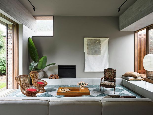 A refined contemporary living room with a low white fireplace wall, grey plaster walls, exposed concrete ceiling beams, expansive timber-framed glazing, and a built-in window seat. A large sectional sofa faces a timber coffee table, woven lounge chairs, patterned rug, sculptural lighting, and restrained artwork, creating a calm blend of modern architecture and eclectic vintage furnishings.